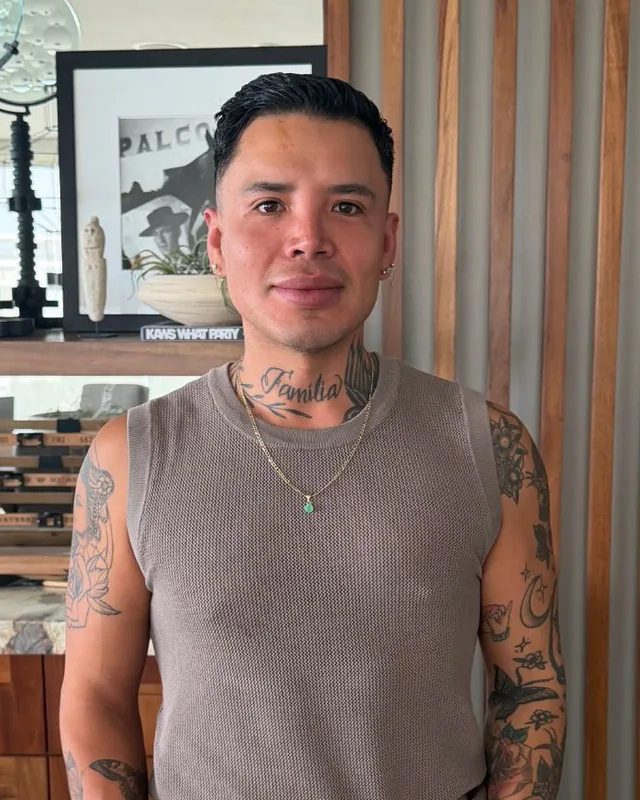Photo of Enrique Meza smiling in a sleeveless knit top, with a Familia tattoo across his neck.